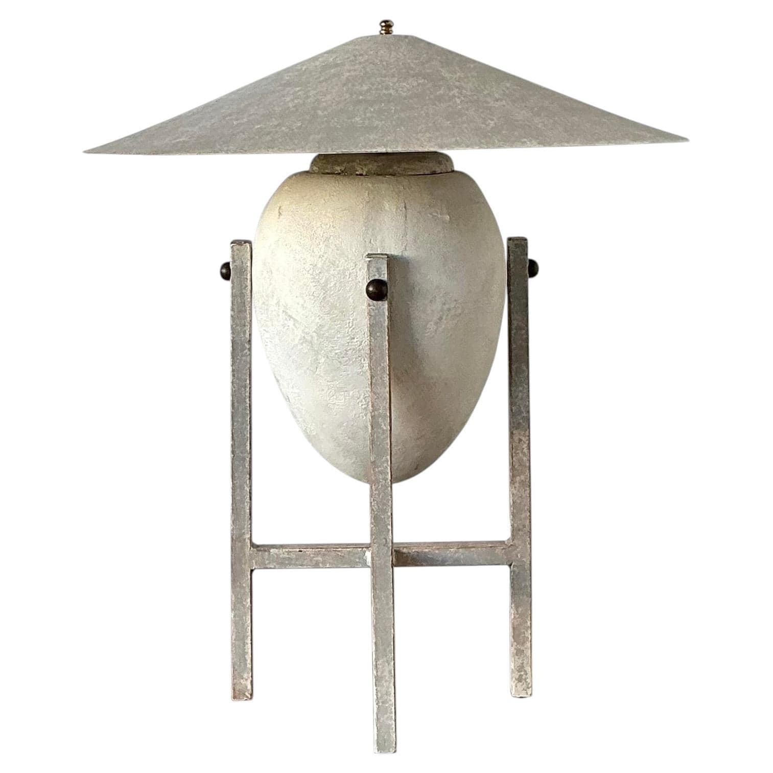 A modernist table lamp featuring an egg-shaped or urn-like body crafted from a textured, light-colored material, likely ceramic or concrete, held within an H-frame metal base. The lamp is topped with a wide, conical metal shade, giving it a distinct brutalist or industrial aesthetic. A small finial adorns the top of the shade. — 3D model