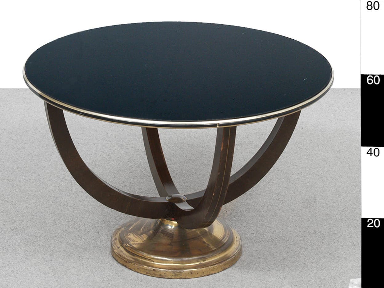 A round side table with a dark, possibly glass or lacquered top, edged with a thin metallic trim. The base features a pedestal-like structure made of brass or a similar metal, supporting curved wooden elements that converge towards the center, creating an open, sculptural design. The wooden legs have a darker finish with visible wood grain and some lighter accents. — 3D model