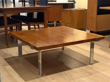 A square coffee table featuring a wooden top and four recessed metal legs. The tabletop has a polished, reddish-brown finish, suggesting a teak or walnut construction. The legs are made of polished chrome or stainless steel, with a rectangular profile, and are integrated into the corners of the table, giving it a minimalist and sleek appearance. The table exhibits characteristics of Mid-Century Modern design. — 3D model