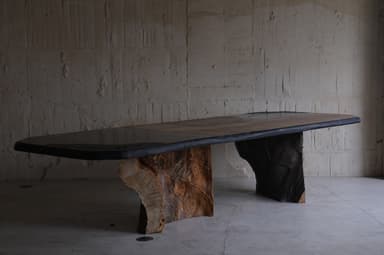 A large dining table with an irregular, organic-shaped top and two substantial, textured bases that resemble natural rock formations or tree trunks. The tabletop appears to be made of multiple panels, possibly wood or stone, with a dark, possibly metal or resin, edge banding. The bases are dark brown and have a rough, sculpted texture, giving the impression of raw, natural elements. — 3D model