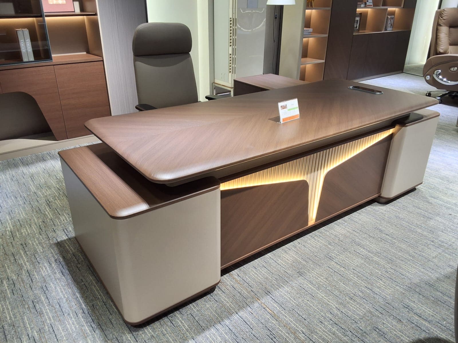 An executive desk featuring a layered, modern design with a dominant wood veneer surface, possibly walnut, showcasing a distinctive herringbone pattern on the main desktop. The desk incorporates integrated LED lighting along its front fascia, creating a warm glow. Side panels are upholstered in a contrasting light-colored leatherette or faux leather, with rounded edges. The desk also includes built-in storage or support structures on the sides and appears to have an integrated power/data grommet on the desktop. — 3D model