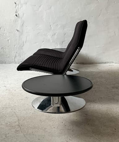 A two-seater sofa with attached side tables. The sofa features black upholstered seats and backrests with a ribbed texture on the backrests. The frame appears to be metal, possibly chrome or stainless steel, with a dark finish on the main horizontal support. Each seat has a cantilevered base with a circular, flared chrome pedestal. Attached to the horizontal frame on either side are circular, dark-colored side tables with a glossy finish, supported by metal brackets. — 3D model
