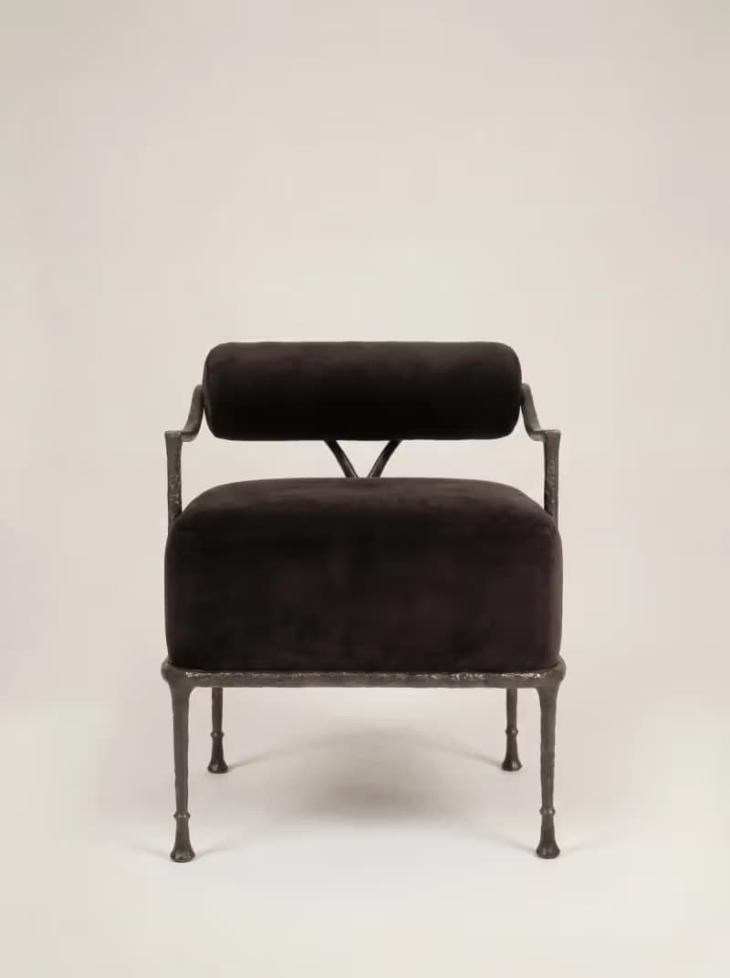 A robust armchair featuring a dark, possibly black or very dark brown, velvet upholstered seat and a cylindrical backrest. The frame is constructed from dark, textured, possibly forged iron or a similar heavy metal, exhibiting a somewhat raw and organic aesthetic. The legs are slender but robust, tapering slightly to small, disc-like feet. The backrest is suspended above the seat, supported by the frame's uprights, with a small V-shaped support element visible at the rear. — 3D model