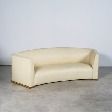 A contemporary curved sofa upholstered in light beige fabric, featuring a continuous back and armrest design, and a recessed plinth base accented with a gold-toned trim. — 3D model