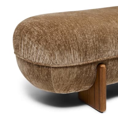 A low-profile, elongated bench with a rounded, upholstered form and solid wood legs. The upholstery features a textured, velvet-like fabric in a neutral tone, exhibiting subtle creasing that follows the rounded shape. The legs are triangular in cross-section and appear to be made of a dark wood with a visible grain. — 3D model