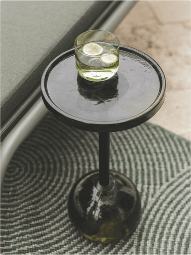 A modern outdoor side table featuring a round, shallow tray-style top, a slender cylindrical pedestal, and a distinctive spherical base. The table is dark in color, likely black or dark gray, with the spherical base appearing to be made of a reflective material such as dark glass or polished plastic. A glass of water with lime slices rests on the tabletop, suggesting its function as a drinks table. The table stands on a paved outdoor surface, next to what appears to be an outdoor lounge chair or sofa. — 3D model
