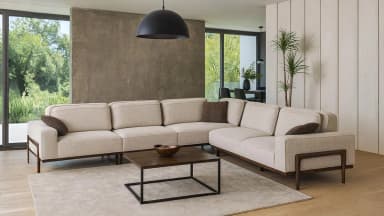A modern, L-shaped sectional sofa with a light beige textured fabric upholstery. It features clean lines, plush seat and back cushions, and a low profile. The sofa is accented with dark wooden armrests and legs that extend slightly beyond the main body, creating a minimalist aesthetic. Two dark brown accent pillows are placed on the sofa. — 3D model