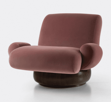 A plush armchair with a rounded silhouette and voluminous cushioning. The seat and backrest are upholstered in a soft, rose-colored velvet fabric. It features integrated armrests that flow seamlessly from the main body of the chair. The chair is supported by a substantial, dark-stained wooden base with a rounded, drum-like shape. — 3D model