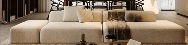 A contemporary modular L-shaped sectional sofa, upholstered in a light beige textile fabric. The sofa features a low-profile design with integrated low backrests and armrests, comprising multiple individual units arranged in an L-configuration. It is accessorized with loose back cushions, a brown patterned throw blanket, and two spherical decorative pillows, suggesting a comfortable and modern aesthetic. — 3D model