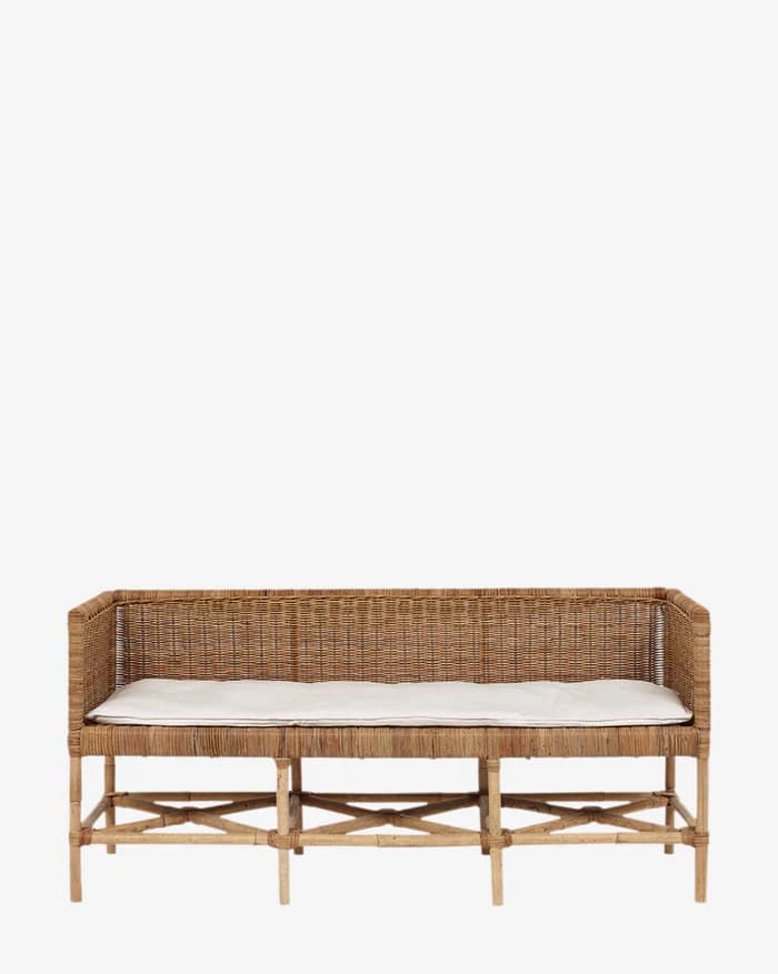 A rustic bench constructed from woven rattan or wicker, featuring a slatted base with X-shaped bracing for support. The bench has a high back and armrests, both made from the same woven material. A plain white cushion is placed on the seat. — 3D model