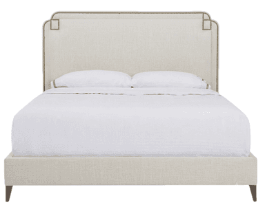 A contemporary upholstered bed featuring a distinctive headboard and a matching upholstered frame. The headboard has a gently curved top edge with subtle cutouts and is adorned with a continuous line of brass or bronze nailhead trim, which also forms decorative square accents at the upper corners. The entire bed frame, including the side rails and footboard, is upholstered in a light, neutral-toned fabric. The bed is supported by four dark, tapered wooden legs and is presented with white bedding. — 3D model