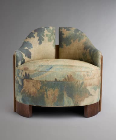 A single armchair with a rounded, upholstered body. The main upholstery features a large-scale floral or botanical pattern in muted greens, blues, and beige tones. The sides of the chair are covered in a contrasting smooth, tan material, possibly suede or a similar textile. The base is a dark wood, possibly walnut or mahogany, with a curved, pedestal-like design. The back of the chair has a subtle wingback element. — 3D model