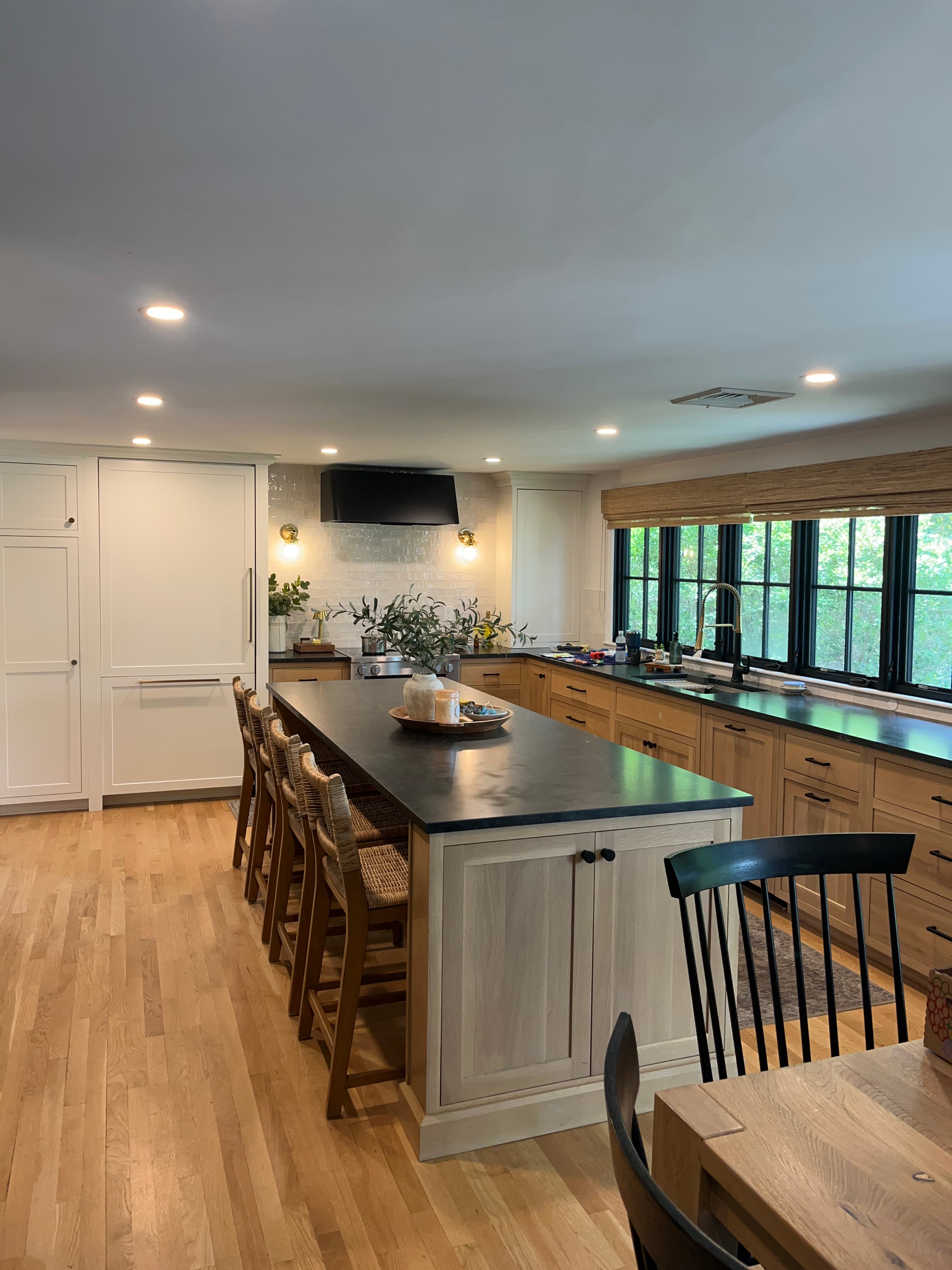 A kitchen island with a light-colored wood base and a dark countertop.  The island features two cabinet doors with dark metal knobs.  Multiple woven-seat stools are positioned around the island. A dark-colored wooden chair is partially visible in the foreground. The island is part of a larger kitchen setting, with additional kitchen cabinets visible in the background. — 3D model