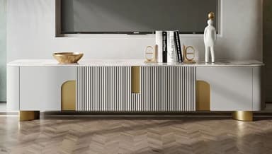 A sleek, contemporary media console featuring a rectangular white marble top with subtle gray veining, set upon a white cabinet body. The cabinet front incorporates vertical fluted detailing and arch-shaped gold accents that serve as visual elements and possibly handles for storage compartments. The corners of the console are rounded, and it rests on short, recessed gold-finished feet. — 3D model