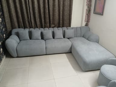 A modern light grey fabric sectional sofa featuring a modular design with a prominent chaise lounge on the right. The backrest and armrests exhibit distinctive channel tufting, creating a plush and rounded aesthetic. Four matching throw pillows are included. The sofa has a low-profile, inviting appearance. — 3D model