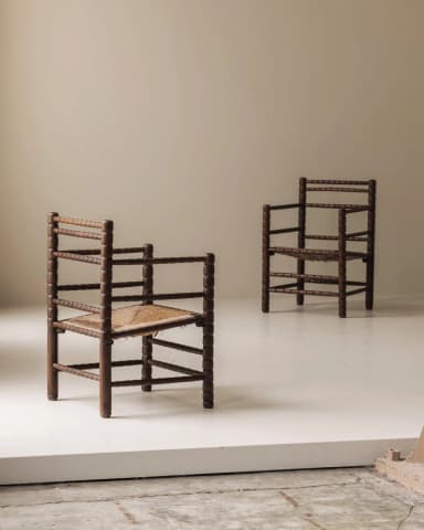 A rustic armchair featuring a dark brown, bobbin-turned wooden frame and a tightly woven rush seat. The chair has a square silhouette with straight legs and armrests, and the backrest is composed of multiple horizontal bobbin-turned spindles. The overall appearance is traditional and handcrafted. — 3D model