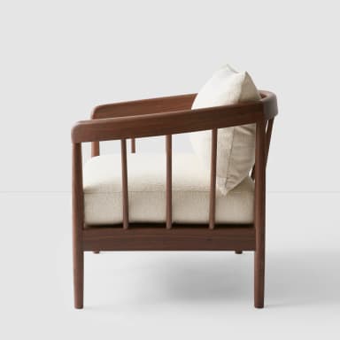 A side profile view of a wooden armchair featuring a curved backrest and slatted sides. The seat and a single throw pillow are upholstered in a textured, light-colored fabric. — 3D model
