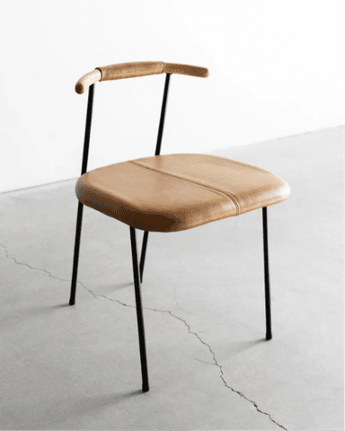 A contemporary dining chair featuring a tan saddle leather upholstered seat with a central stitch line, set on a slender black metal frame. The backrest consists of two upright black metal rods supporting a gently curved wooden bar, partially wrapped in matching saddle leather, which also functions as a minimal armrest. The overall design is minimalist and clean-lined. — 3D model