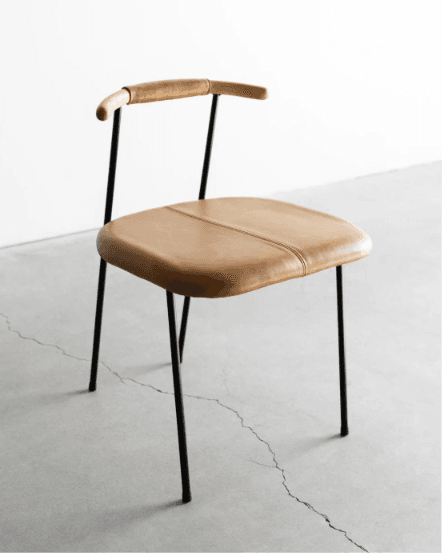 A contemporary dining chair featuring a tan saddle leather upholstered seat with a central stitch line, set on a slender black metal frame. The backrest consists of two upright black metal rods supporting a gently curved wooden bar, partially wrapped in matching saddle leather, which also functions as a minimal armrest. The overall design is minimalist and clean-lined. — 3D model