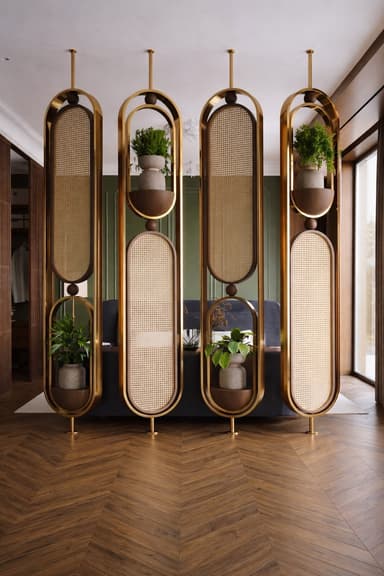 A tall, narrow, wall-mounted display unit featuring an elongated oval frame constructed from brass-toned metal and dark wood. It is divided into three sections: two sections contain semicircular wooden shelves, each holding a potted plant, and the central section features a panel of natural cane webbing. The design incorporates rounded edges and a harmonious blend of materials, suggesting an Art Deco or contemporary aesthetic. — 3D model