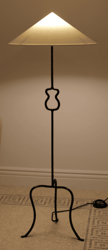 A tall, slender floor lamp featuring a black wrought iron (or similar dark metal) stand and a conical, off-white shade. The stem includes a distinctive curvilinear decorative element in its mid-section. The lamp is supported by a splayed, three-legged tripod base with curved feet. — 3D model