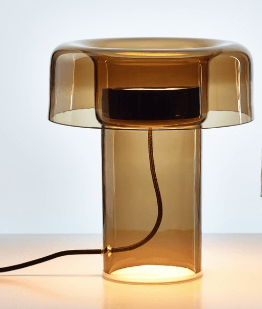 A modern table lamp constructed primarily from smoked amber glass. The design features a cylindrical base that tapers slightly upwards, topped by a mushroom-shaped diffuser. The diffuser consists of a wider, flatter glass element that extends outwards and over a smaller, darker cylindrical component. A fabric-covered power cord exits the base. The lamp emits a warm, diffused light from its base and the area beneath the diffuser. — 3D model