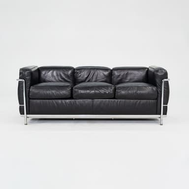 A three-seater sofa characterized by its boxy, geometric silhouette and exposed tubular chrome-plated steel frame. It features black leather upholstery on separate, plump cushions for the seat, back, and arms. The cushions appear to be removable and are designed to sit within the sturdy metal structure. The leather shows signs of use, such as creasing, indicating a well-loved piece. — 3D model