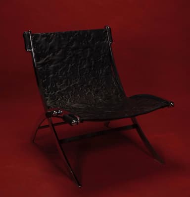 A modern lounge chair with a striking contemporary design. The chair features a polished chrome metal frame with a distinctive angular and curved structure, supporting a seat and backrest upholstered in black material with a textured, crinkled finish. The armrests consist of cylindrical black elements secured by chrome fixtures. — 3D model