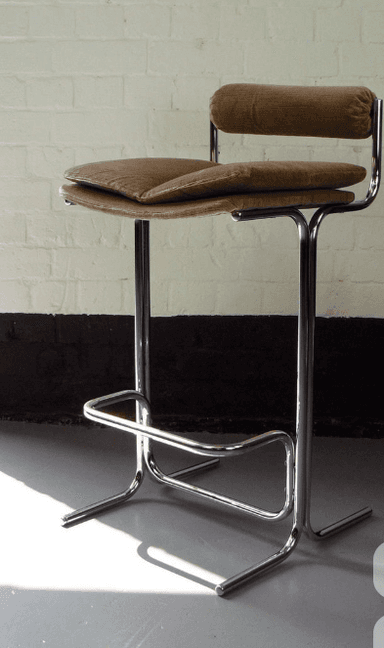 A bar stool with a chrome-plated metal frame and a cushioned seat and backrest. The seat and backrest are upholstered in a ribbed fabric with a light brown or tan color. The frame features a tubular design with a footrest. The stool has a modern and minimalist aesthetic. — 3D model