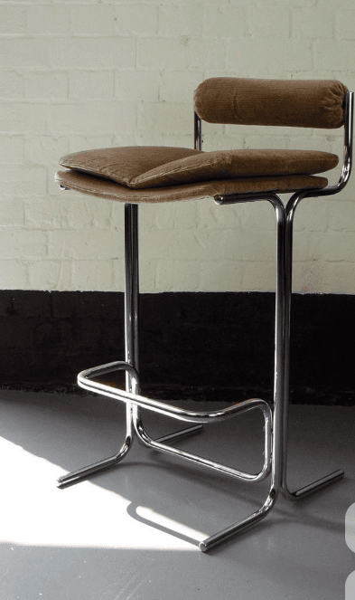 A bar stool with a chrome-plated metal frame and a cushioned seat and backrest. The seat and backrest are upholstered in a ribbed fabric with a light brown or tan color. The frame features a tubular design with a footrest. The stool has a modern and minimalist aesthetic. — 3D model