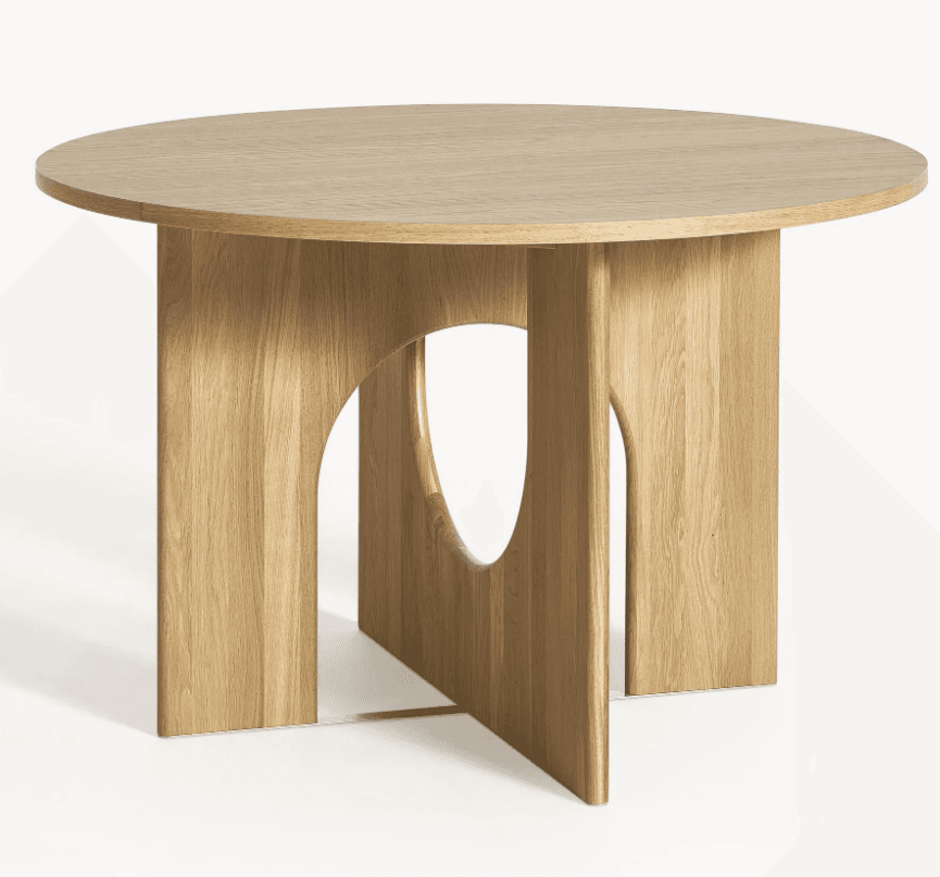A round dining table features a smooth, circular tabletop crafted from light-toned wood with visible grain. It is supported by a distinctive sculptural pedestal base, which is constructed from four broad, vertical wooden panels. These panels are arranged symmetrically, forming a central cruciform structure at the bottom, and each incorporates a large, arcing cutout that creates rounded negative spaces, lending an airy and contemporary aesthetic. — 3D model