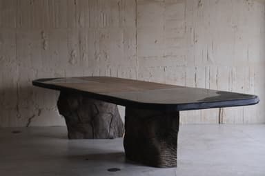 A large dining table with an irregular, organic-shaped top and two substantial, textured bases that resemble natural rock formations or tree trunks. The tabletop appears to be made of multiple panels, possibly wood or stone, with a dark, possibly metal or resin, edge banding. The bases are dark brown and have a rough, sculpted texture, giving the impression of raw, natural elements. — 3D model