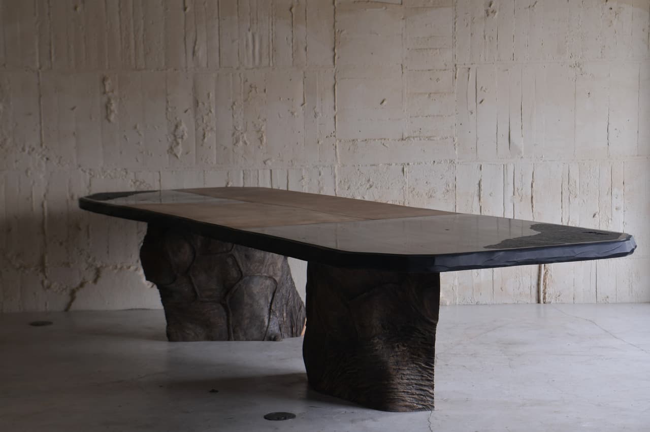 A large dining table with an irregular, organic-shaped top and two substantial, textured bases that resemble natural rock formations or tree trunks. The tabletop appears to be made of multiple panels, possibly wood or stone, with a dark, possibly metal or resin, edge banding. The bases are dark brown and have a rough, sculpted texture, giving the impression of raw, natural elements. — 3D model