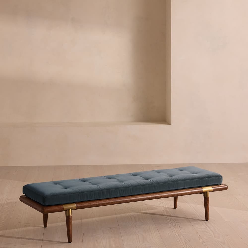 A long, upholstered bench with a wooden frame. The seat is tufted and covered in a dark, possibly blue or gray, fabric. The frame appears to be made of dark wood and features four tapered legs. Brass accents are visible at the connection points between the legs and the frame. The design suggests a mid-century modern or contemporary style. — 3D model