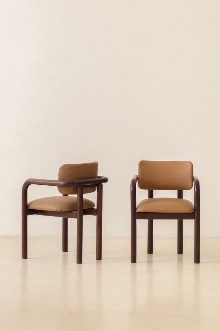 Two armchairs with wooden frames and upholstered seats and backrests. The frames have a rounded, tubular appearance. The seat and backrest are padded and covered in a light brown material, possibly leather or faux leather. The chair on the left has a slightly more extended armrest that curves upwards to form a small support, while the chair on the right has a standard armrest. The legs are straight and columnar. — 3D model