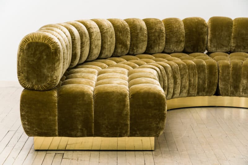 A curved sectional sofa with a channeled upholstery design. The sofa features deep, rounded cushions that run along its entire length, creating a tufted appearance. It is supported by a low-profile, possibly metallic, base with a polished finish. — 3D model