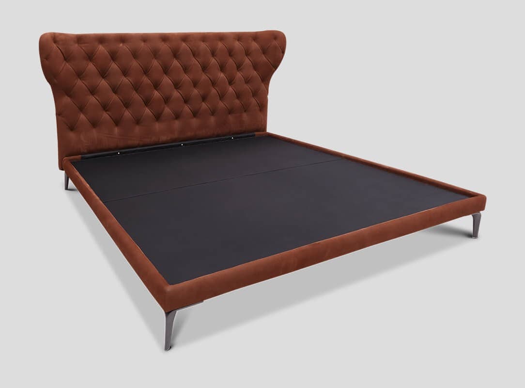 A tufted bed frame with a winged headboard, upholstered in a rust-colored fabric. The headboard features a diamond button-tufted pattern. The frame is designed to support a mattress and has minimal side rails. It rests on four slender, angled legs made of metal. — 3D model