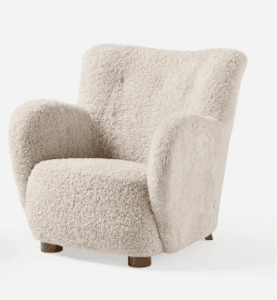 A plush armchair upholstered in a textured, sheepskin-like fabric. It features a high, slightly curved backrest, generously padded arms that flow into the seat, and a rounded seat cushion. The chair is supported by four short, cylindrical wooden legs, visible at the base. — 3D model