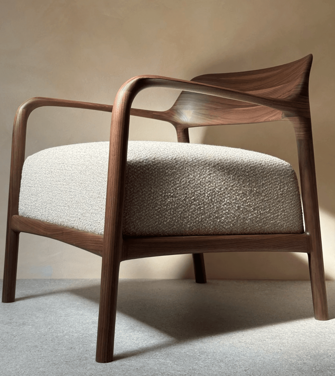 A contemporary armchair featuring a dark-stained walnut wooden frame with smoothly curved lines, integrating armrests and a sculpted backrest. The seat cushion is upholstered in a light, textured woven fabric, likely bouclé or a similar blend, providing a neutral contrast to the warm wood. — 3D model
