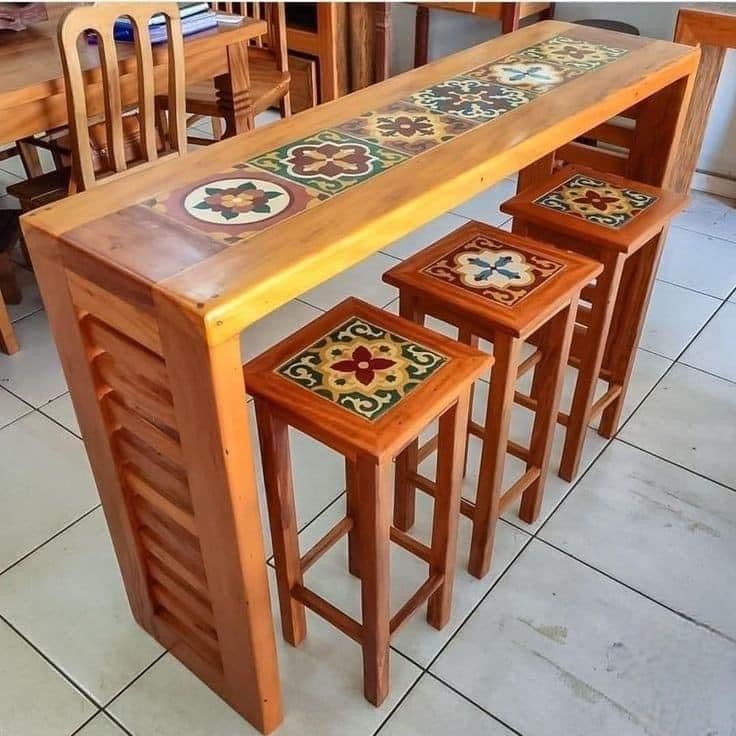 The image features a rustic-style bar table and three matching bar stools, all crafted from light-toned wood with decorative ceramic tile inlays. The table has a long, narrow top with a central runner of tiles, and one visible leg featuring a slatted design. The three stools are tall, square, and also have matching tiled tops, completing a cohesive set. — 3D model