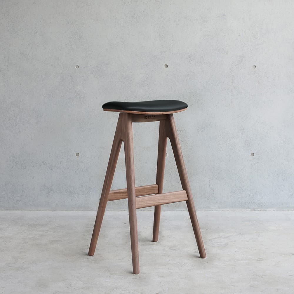 A modern bar stool with a saddle-shaped seat and a wooden frame. The seat is upholstered in black material, possibly leather or faux leather. The frame consists of four angled legs connected by horizontal support bars. The overall design is minimalist and Scandinavian-inspired. — 3D model