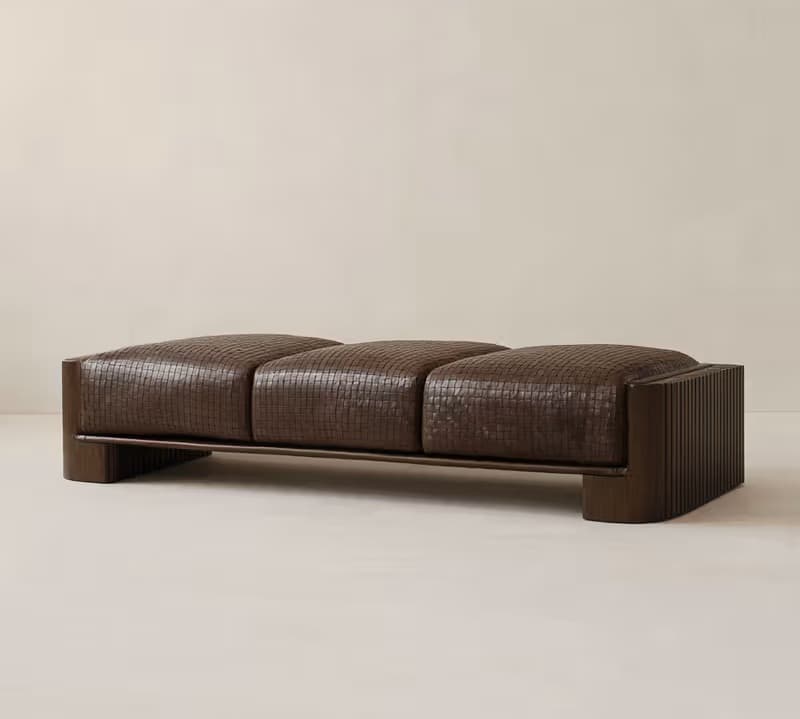 A long, upholstered bench with three distinct seat cushions. The upholstery appears to be a woven leather or textured fabric in a dark brown hue. The frame is made of wood, with a dark finish. The sides of the bench feature a slatted design. — 3D model