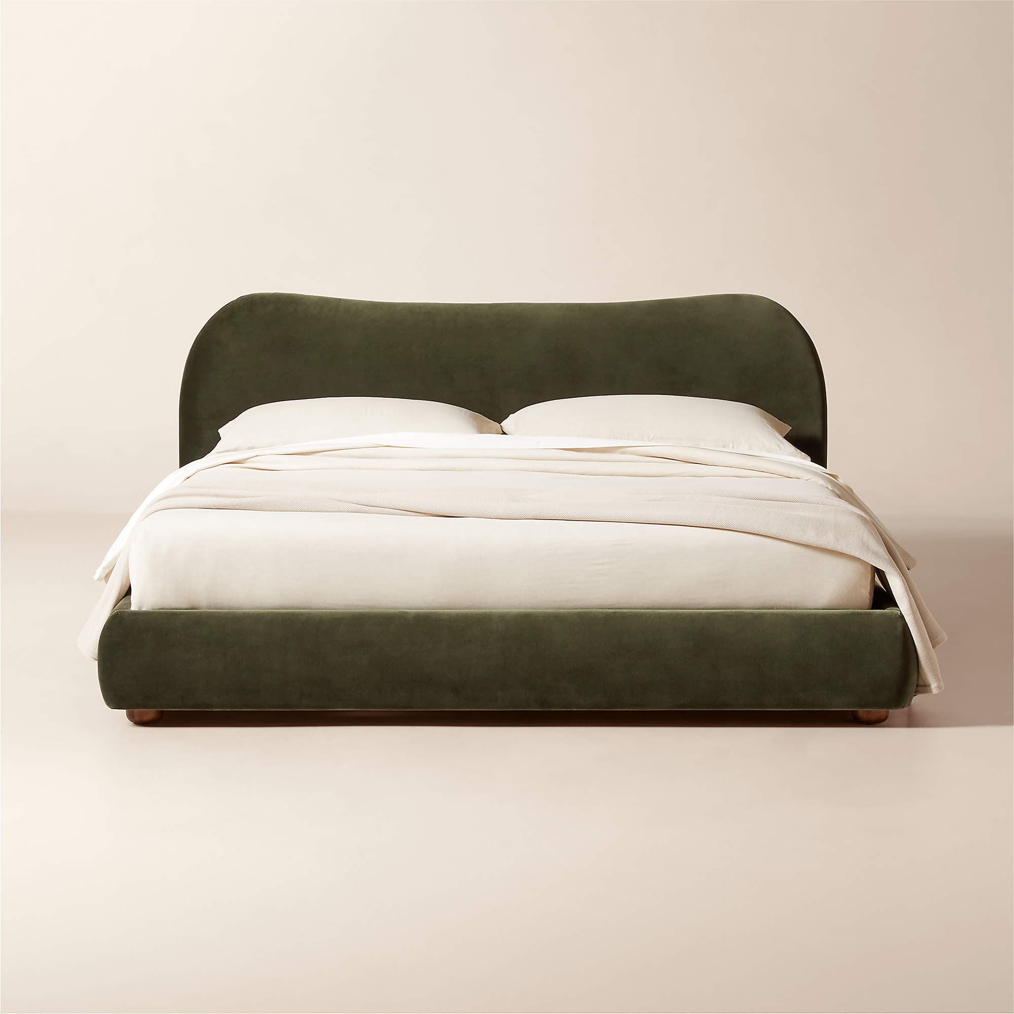 A upholstered bed frame with a curved headboard and a low-profile base. The bed is shown with off-white bedding, including two pillows and a draped comforter. The upholstery appears to be a soft fabric, possibly velvet, in a deep olive green color. The base has a subtle wooden trim visible at the bottom. — 3D model