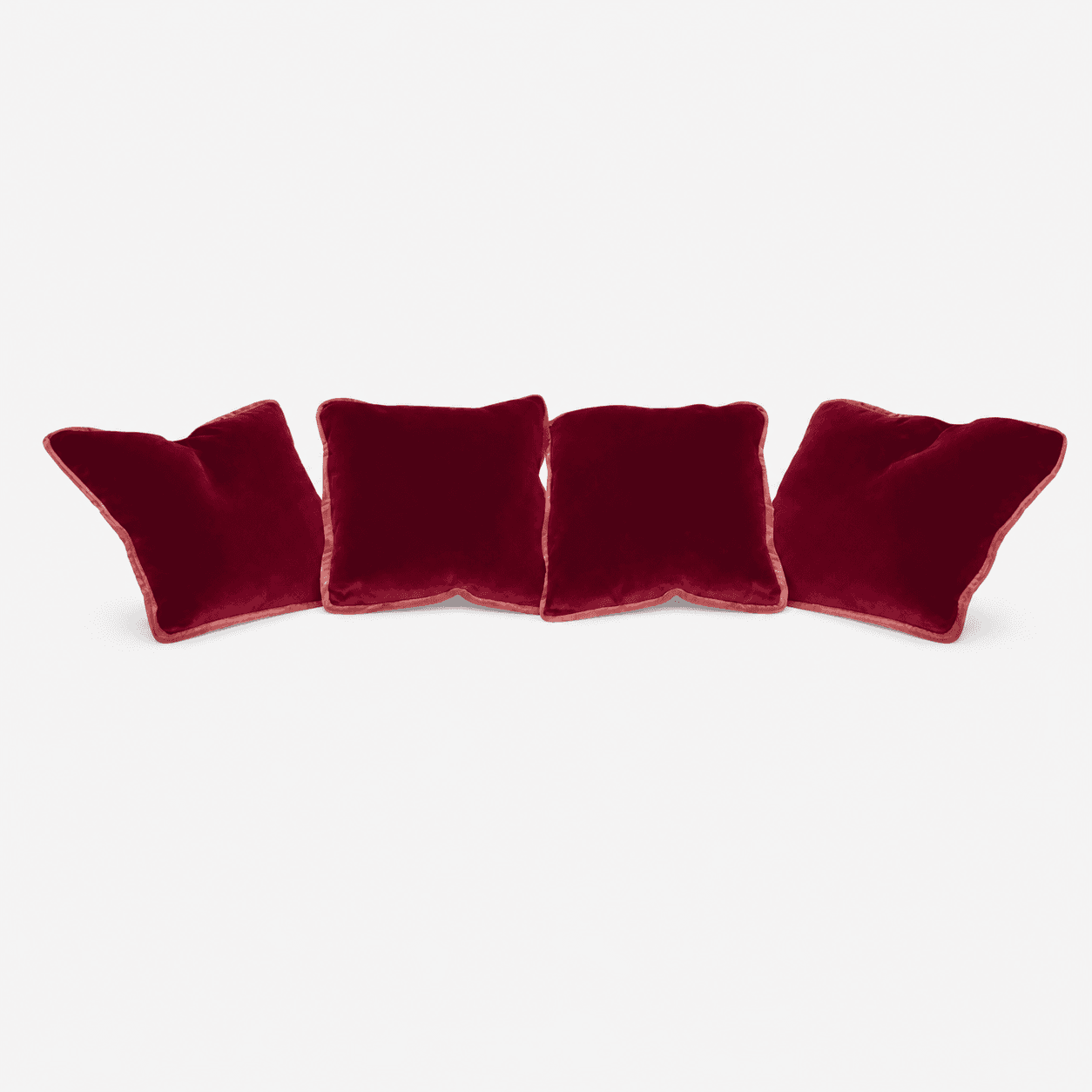 A photograph displays a set of four square-shaped throw pillows, each upholstered in a rich, deep red velvet fabric. The pillows are arranged in a horizontal line and feature a distinct piping along all edges, rendered in a slightly lighter, complementary shade of red. The velvet material exhibits a subtle sheen, characteristic of the fabric. — 3D model