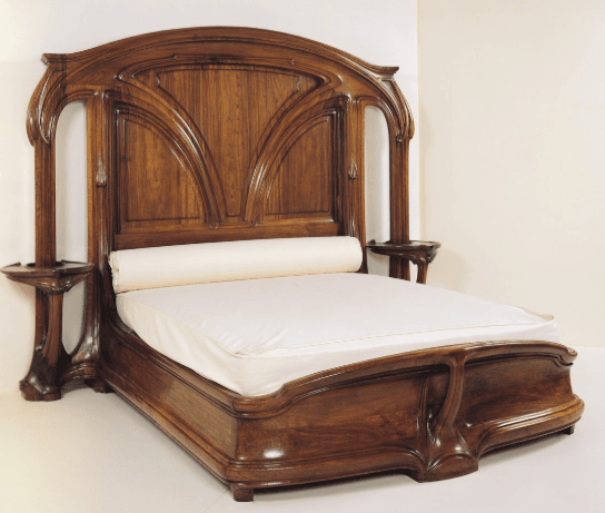 A large, ornate wooden bed frame with a high headboard featuring intricate carved detailing resembling arched windows or leaves. The bed includes two attached nightstands on either side, extending from the headboard structure. The bed frame itself is also carved with flowing lines and has a low footboard. A rolled headrest and a white mattress are present. — 3D model