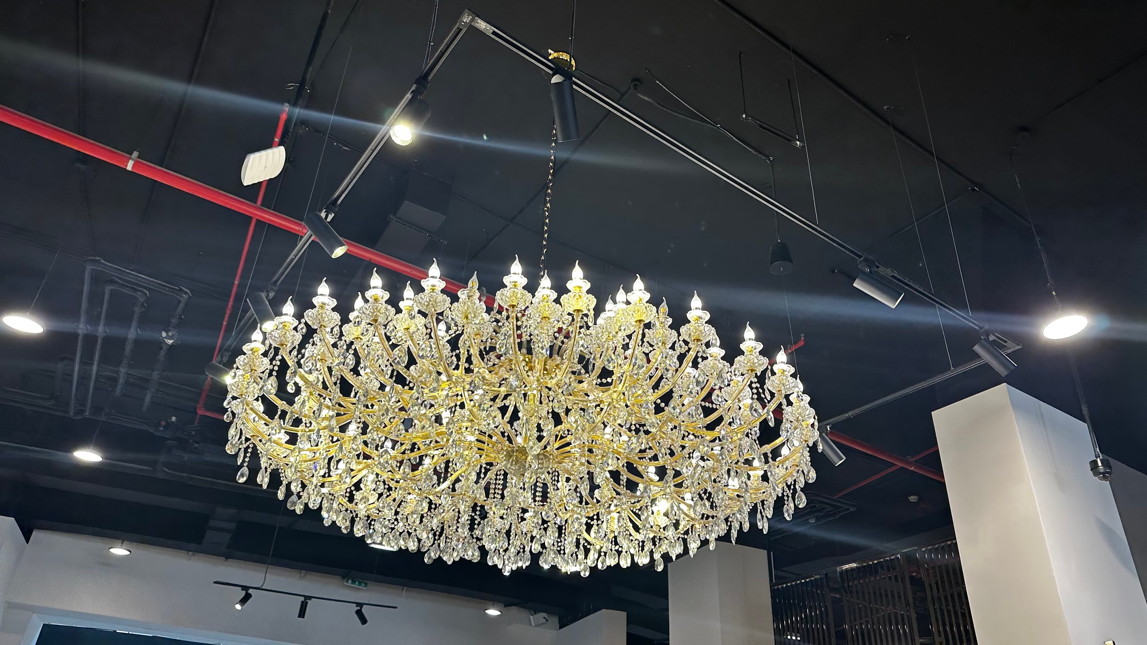 A large, multi-tiered chandelier with a gold-toned metal frame adorned with numerous clear crystal prisms. The chandelier features multiple arms extending outwards, each holding a light source, and cascading crystal elements that refract light. It is suspended against a black background. — 3D model