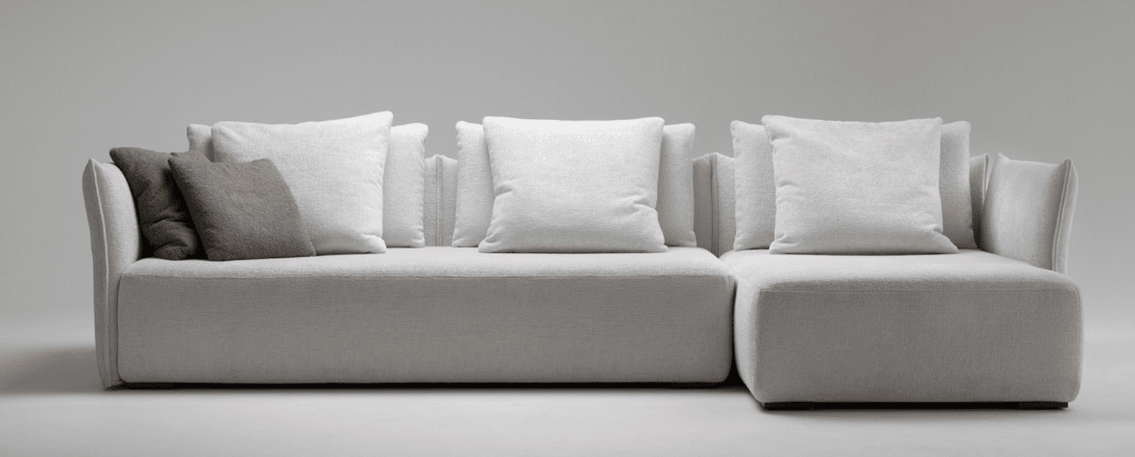 A modern, modular sectional sofa with a light grey upholstery. The sofa features deep, plush seating and a low profile. It is accompanied by several large, square back cushions and two accent pillows, one in a neutral taupe color and another in a similar light grey. The armrests are integrated into the main structure and are slightly sloped. — 3D model