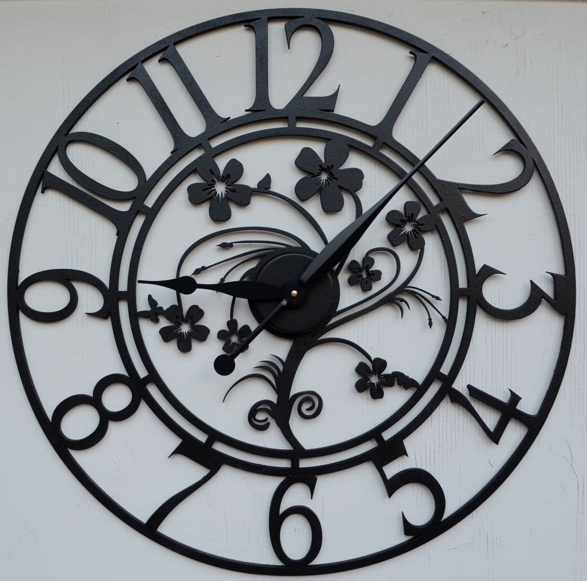 This is an ornate circular wall clock crafted from black metal, featuring an open-face design. The clock face is defined by a large outer ring with cut-out Roman numerals for 12, 11, 10, 9, 8, 7, and Arabic numerals for 6, 5, 4, 3, 2, 1. The central mechanism is surrounded by intricate floral and scrollwork motifs, also cut from black metal. It has black hour and minute hands. — 3D model