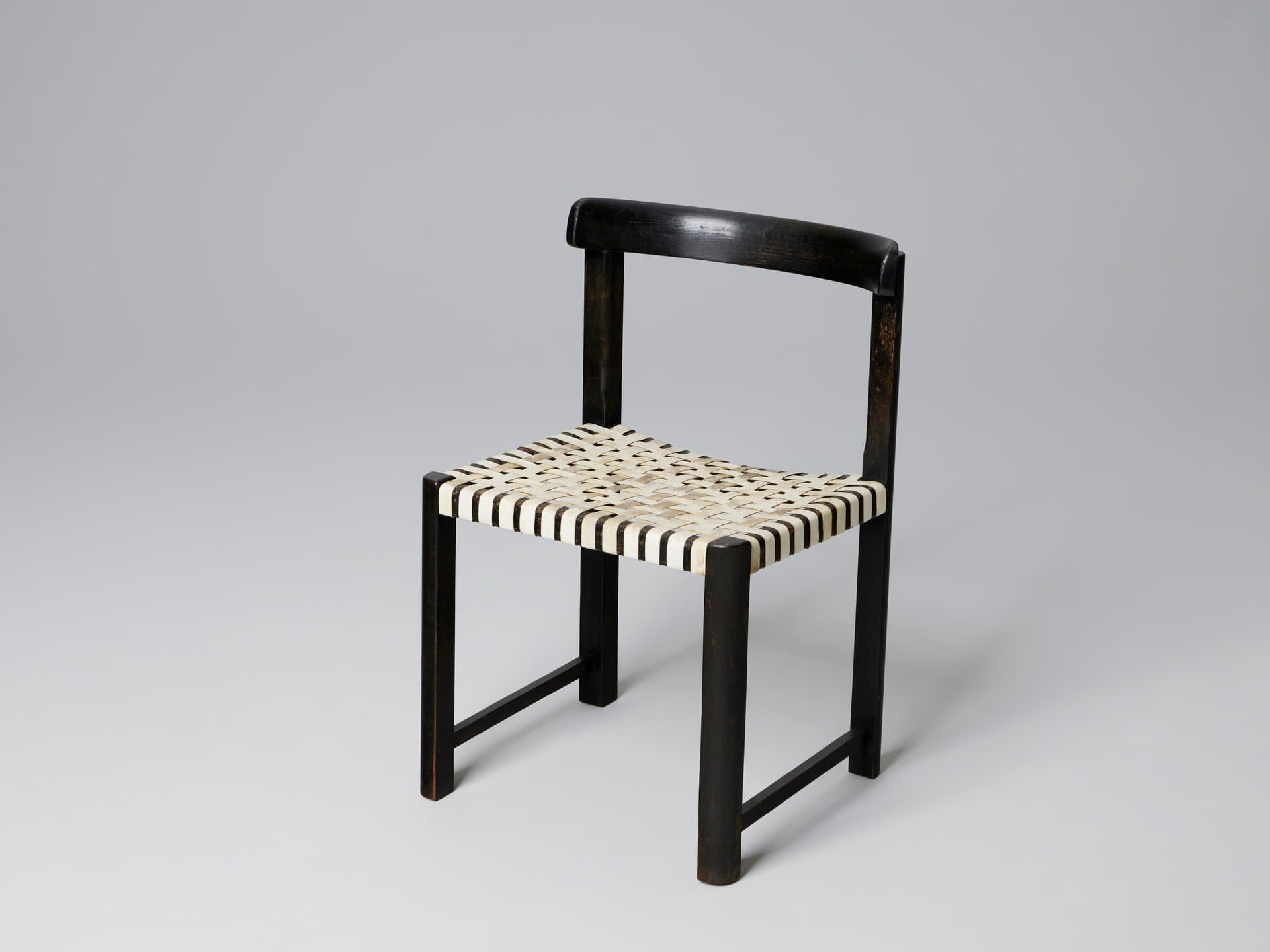 A dark-colored chair with a curved backrest and a woven seat. The frame appears to be constructed from wood or a similar material, with a matte finish. The seat is made of interlaced straps, creating a textured pattern. The legs are straight and set at an angle, connected by a lower stretcher bar on each side. — 3D model