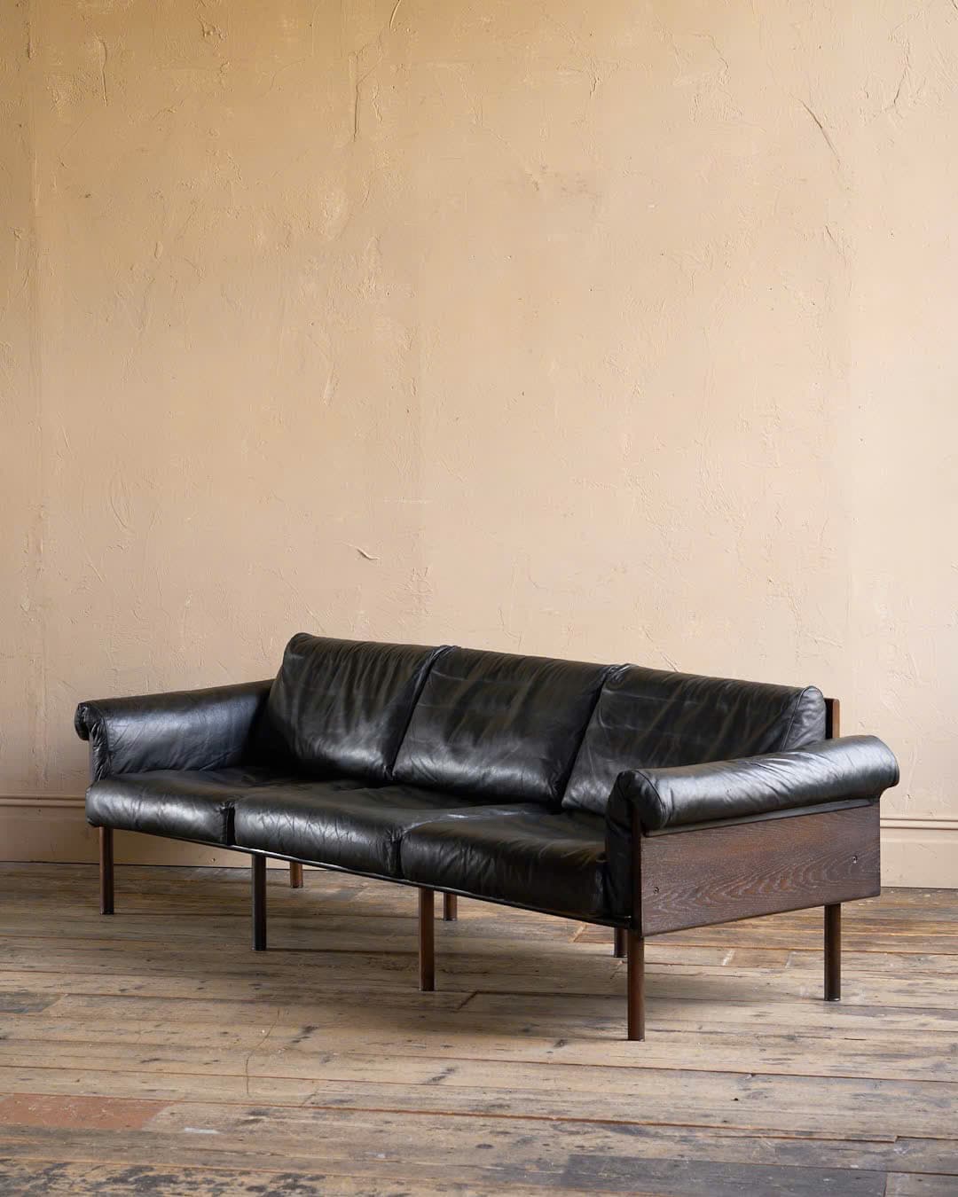 A three-seater sofa upholstered in aged black leather, featuring an exposed dark wood frame that forms the back and side panels, with cylindrical wooden legs. The armrests are rolled and integrated with the leather upholstery. — 3D model